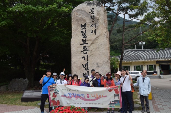 시각장애인 봄 나들이에 참석한 모든 분들이 문경새재 기념비 앞에서 현수막을 들고 단체사진을 찍고 있는 모습