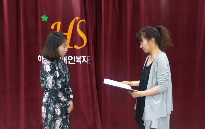 복지관 강당 앞에서 교육치료팀 신입직원 신지영 선생님이 서혜미 국장님으로부터 입명장을 전달받고 있는 모습