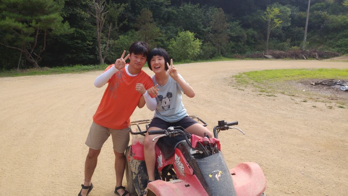 한명은 4륜 바이크에 타고, 한명은 서서 손에 브이를 하며 기념사진을 찍고 있는 모습