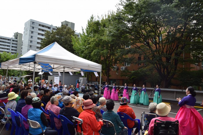 복지관 앞마당에서 한복을 입은 여성들이 노래를 부르고 있는 모습과 관중들이 무대를 보며 호응하고 있는 모습