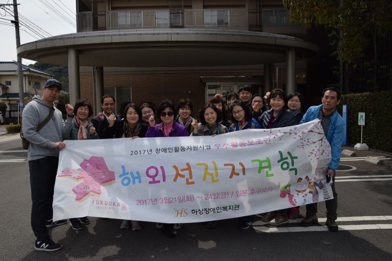 일본 태양의집 앞에서 복지관 직원과 활동보조인이 현수막을 들고 단체사진을 찍고 있음